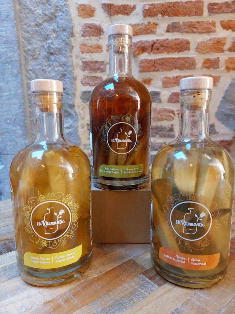 Trois bouteilles de rhums arrangés So'Rhumantic sur une table en bois, avec des étiquettes indiquant Ananas Victoria & Vanille Bourbon, Poire Conférence & Fève de Cacao, et Mangue & Fruit de la Passion.