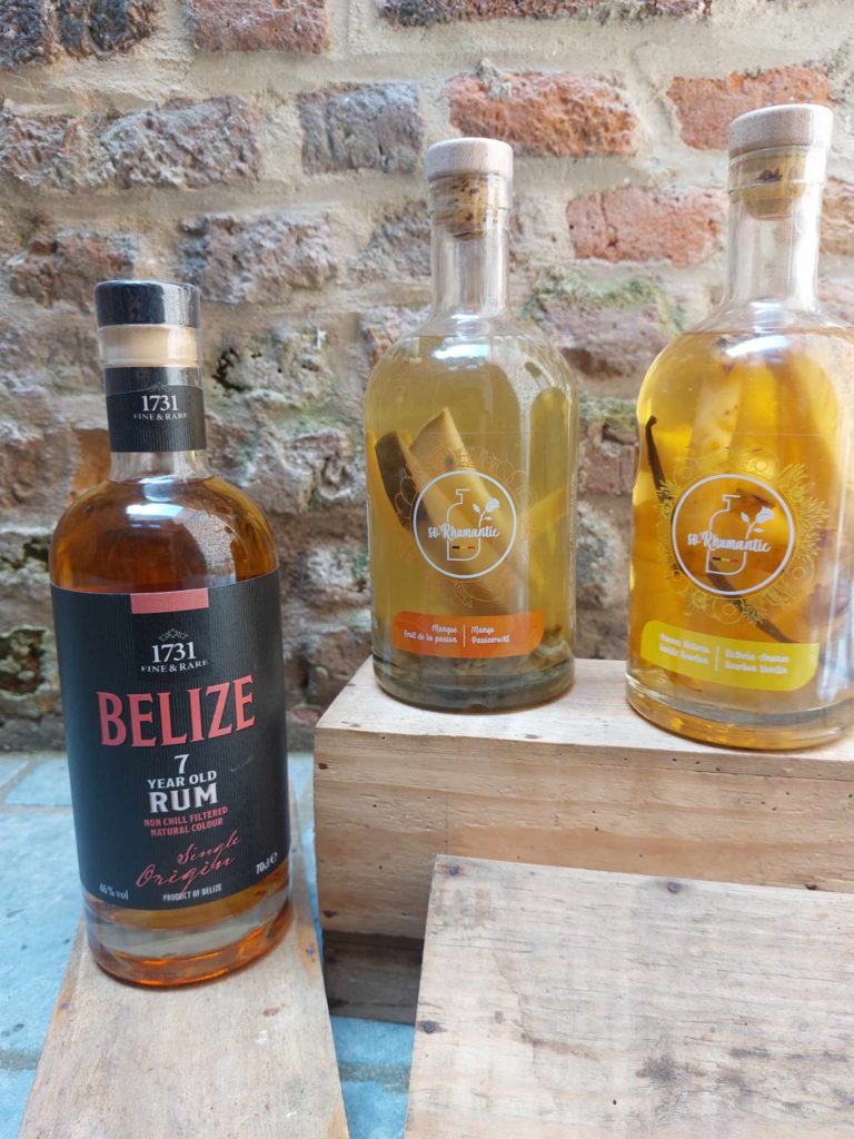 Trois bouteilles d'alcool, dont un rhum Belize 7 ans et deux bouteilles de rhum So'Rhumantic, posées sur des caisses en bois avec un mur en briques en arrière-plan.