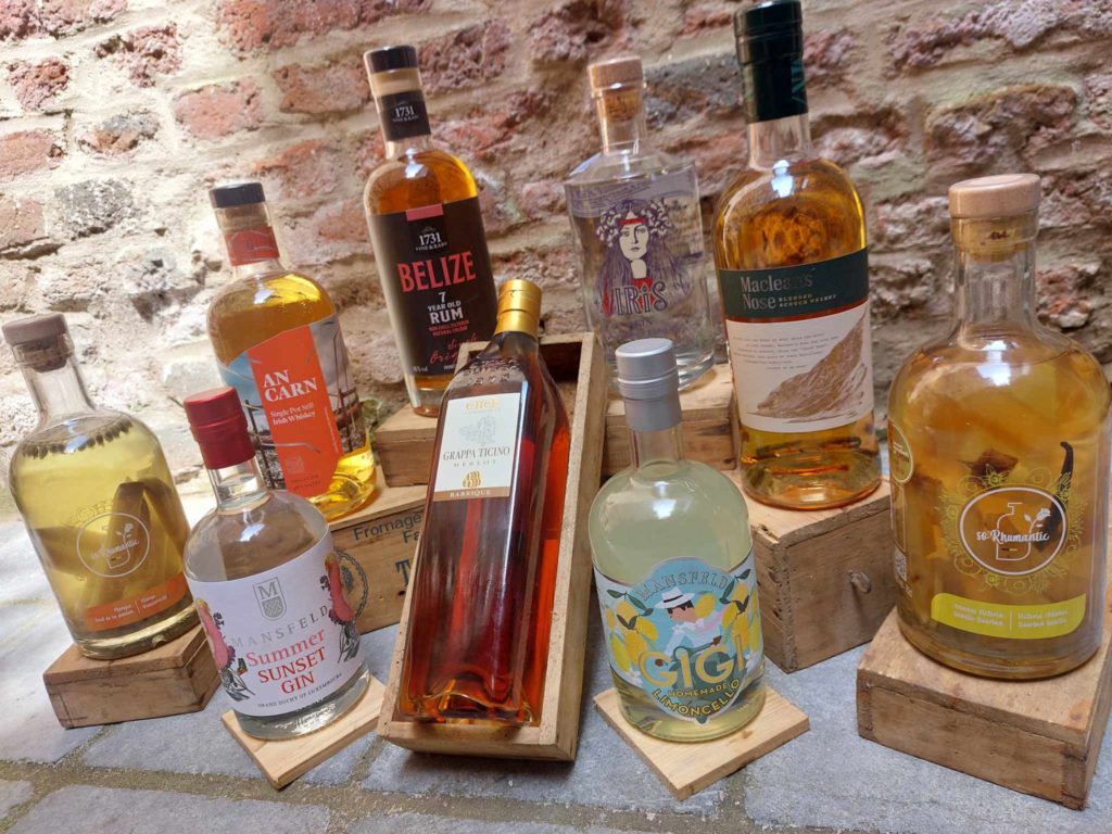 Plusieurs bouteilles d'alcool comprenant des whiskies, gins, rhums et liqueurs, disposées sur des caisses en bois avec un mur en briques en arrière-plan.