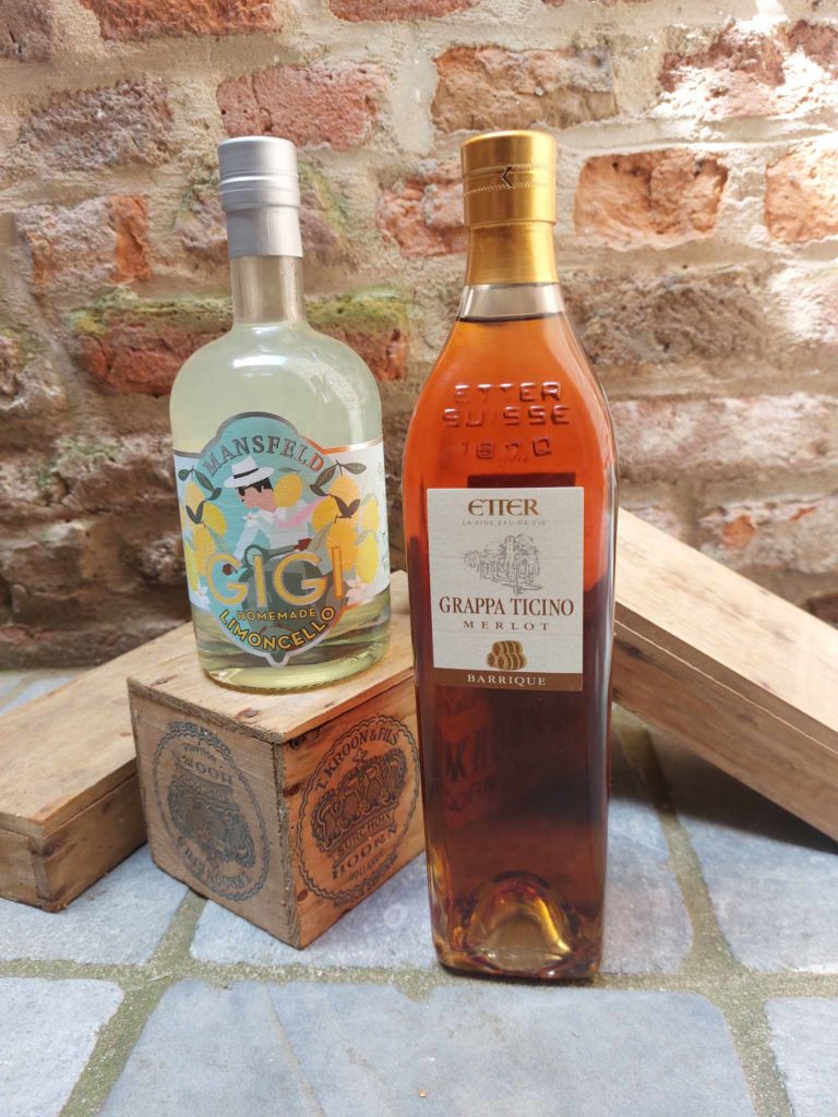 Deux bouteilles d'alcool, Mansfeld Homemade Limoncello et Etter Grappa Ticino Merlot, posées sur des caisses en bois avec un mur en briques en arrière-plan.