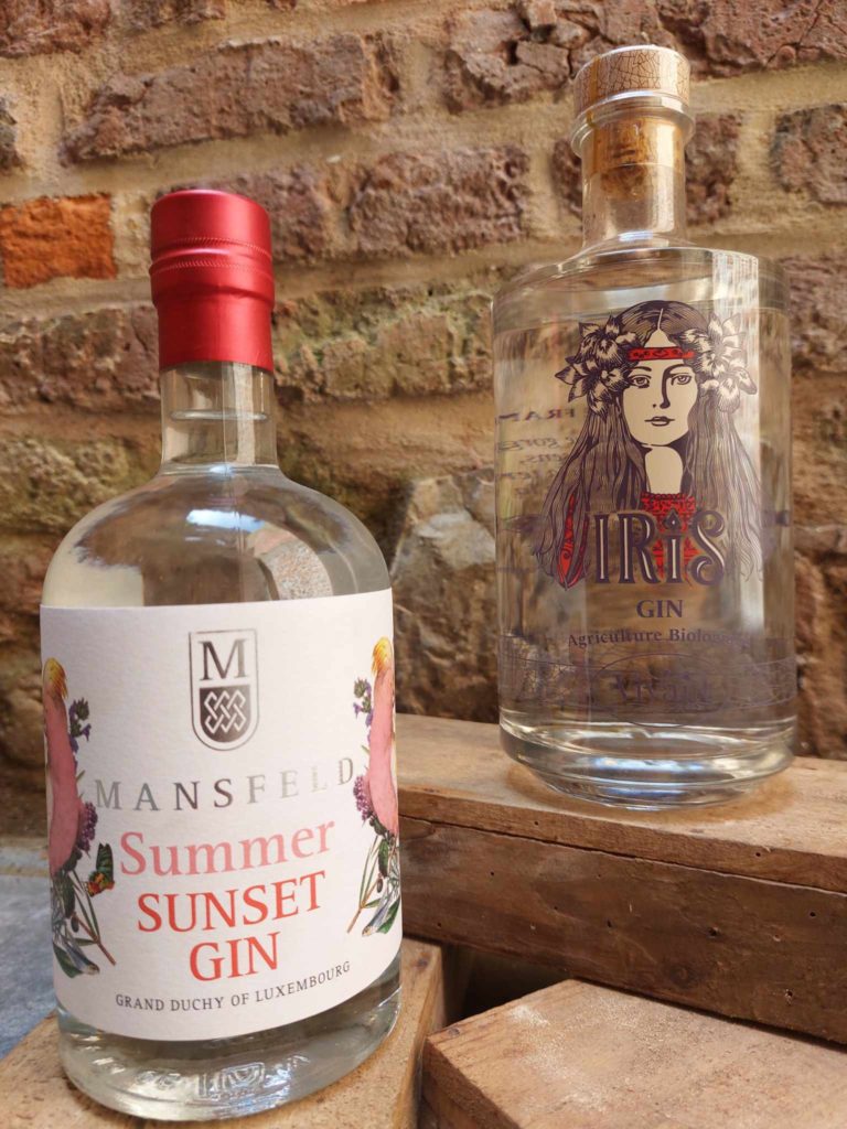 Deux bouteilles de gin, Mansfeld Summer Sunset Gin et Vivant Gin Iris, posées sur des caisses en bois avec un mur en briques en arrière-plan.